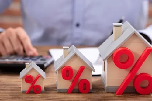 Três maquetes de casas em tamanhos crescentes com símbolos percentuais vermelhos à frente, sobre uma mesa, enquanto uma pessoa utiliza uma calculadora ao fundo, representando taxas de juro e spread no crédito habitação.